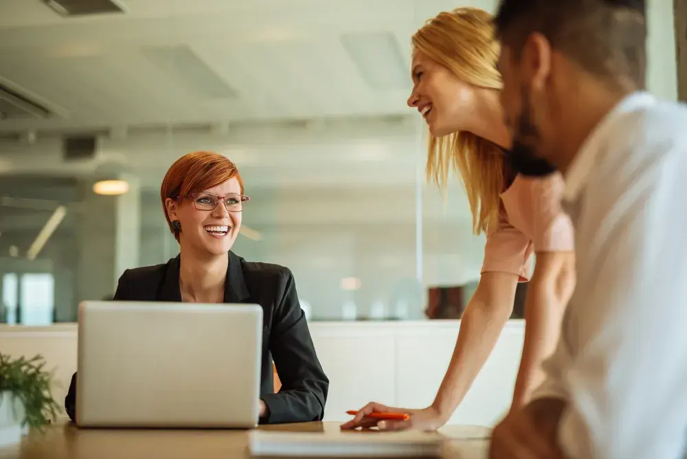 Three colleagues in a modern office setting, smiling and having a conversation around a laptop.