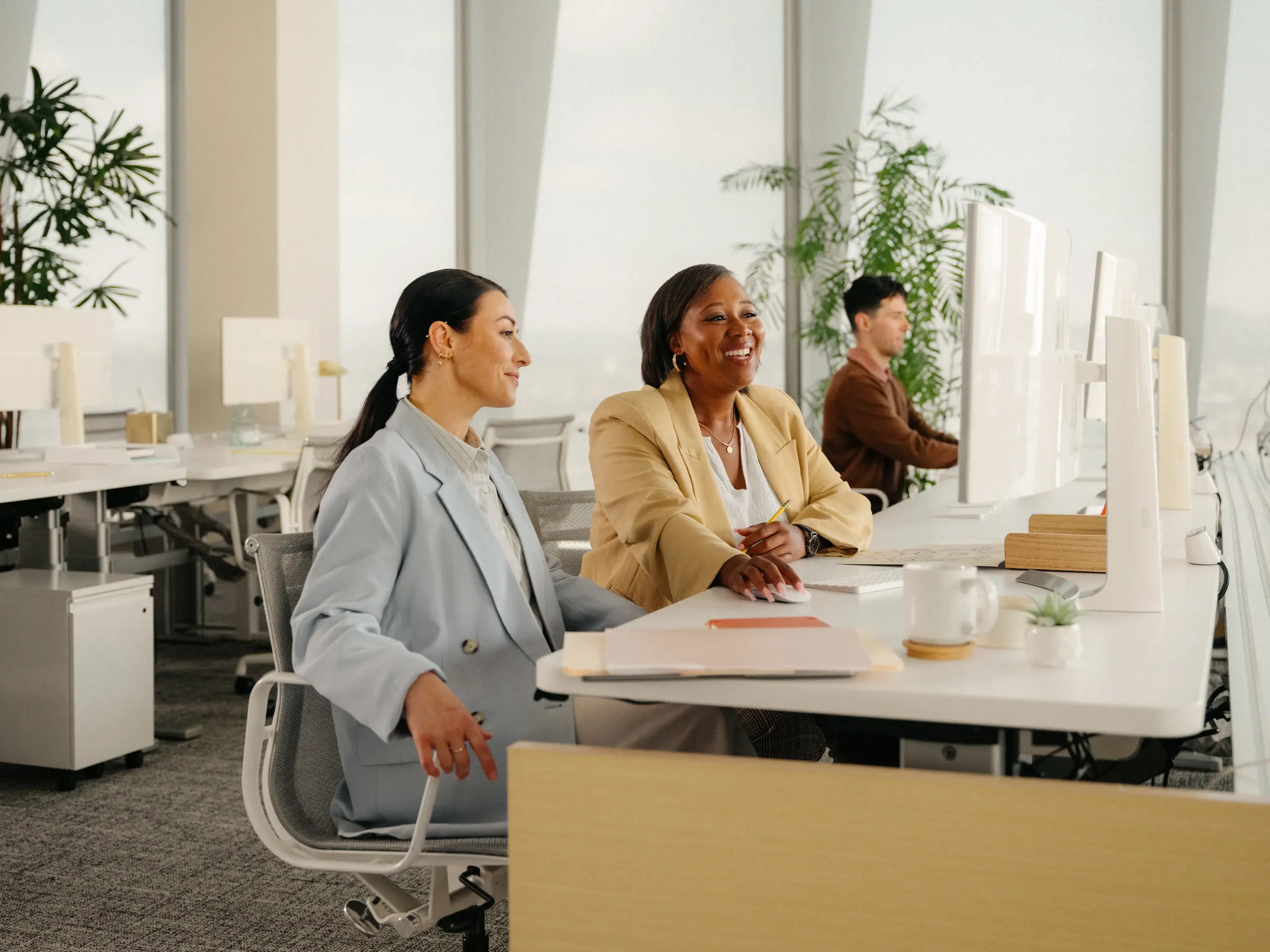 Gemini said Two female professionals collaborating at a desk in a bright, modern office. One woman sits while the other leans in, smiling and using the computer mouse, with a male colleague working in the background.