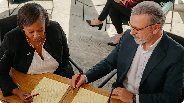 Twee professionals, een man en een vrouw, zitten tijdens een vergadering zij aan zij aan een houten tafel. Ze kijken allebei naar beneden en schrijven op gele notitieblokken, en lijken samen te werken aan hun notities.
